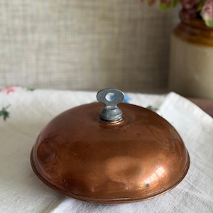 May include: A round, polished copper lid with a smooth, domed surface and a central metallic handle featuring a small circular opening at the top. The copper has a warm, reflective finish with subtle variations in tone across its curved exterior.