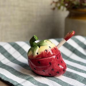 May include: A ceramic strawberry-shaped jar with a removable lid featuring a green stem and a matching small spoon. The jar is painted in shades of red with black seed details and a cream-coloured top section, designed for holding condiments or sugar.