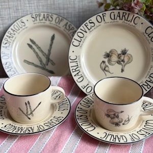 May include: A set of cream-coloured ceramic tableware featuring two plates, two saucers, and two cups. The items are decorated with black botanical illustrations of asparagus and garlic, with the text "SPEARS OF ASPARAGUS" and "CLOVES OF GARLIC" printed along the rims.