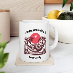 May include: White ceramic mug with a brown sloth illustration in a hammock with palm trees and a red sun. The text "I'll Get Around To It Eventually" is printed on the mug.