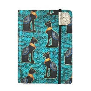 May include: A notebook with a teal background and a repeating pattern of black cat figures with gold and blue accents. The notebook has a black elastic band and a silver-colored closure.