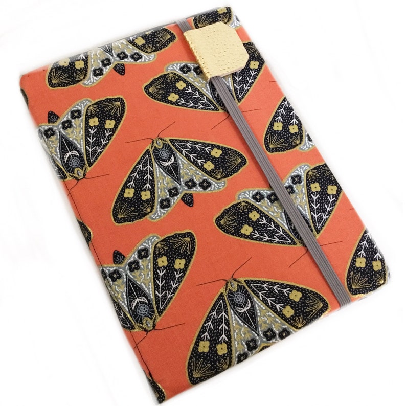 Pode incluir: Uma capa de caderno de cor laranja coral com um padr&atilde;o de mariposa preto e dourado. A capa tem um fecho el&aacute;stico cinzento e um pequeno marcador amarelo.
