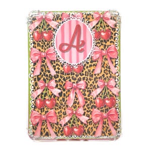 May include: A decorative rectangular case with a leopard print background, pink bows, and cherries. A pink letter "A" is centered on a pink striped background. The case has a white lace-like border and a clear plastic frame.