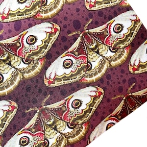 May include: Close-up of a fabric with a repeating pattern of large moth illustrations. The moths have white, brown, red, and yellow wings with a central eye-like design. The background is a dark purple with small, lighter purple dots.