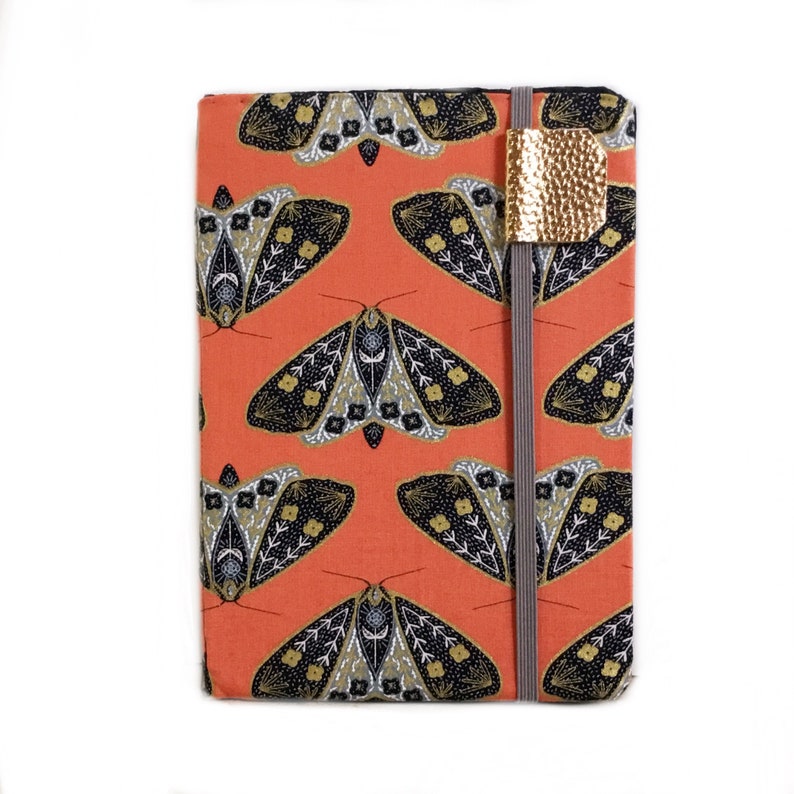 Pode incluir: Uma capa de caderno laranja coral com um padr&atilde;o de mariposa preto e dourado. A capa tem um marcador de metal dourado.