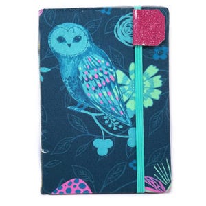 May include: A notebook with a dark blue cover featuring a teal owl illustration with pink accents. The design includes floral and leaf patterns. A pink glittery tag and teal elastic band secure the notebook.
