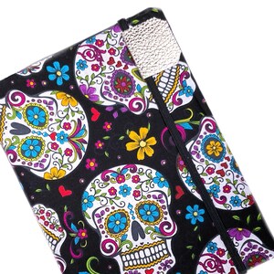 May include: A black journal with a colorful sugar skull design. The cover features multiple sugar skull illustrations with floral and heart accents in white, blue, yellow, and purple. A silver-colored elastic closure and page marker are visible.