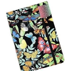 May include: A rectangular journal with a black background covered in colorful butterflies and green leaves. The butterflies are in shades of blue, yellow, orange, and white. A glittery, multi-colored bookmark is attached to the journal with a black elastic band.