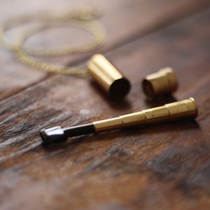 May include: A gold-coloured, miniature telescope pendant with a black eyepiece, lying on a wooden surface. The pendant is partially disassembled, with the cap and a separate cylindrical piece visible. A delicate gold chain is also present.