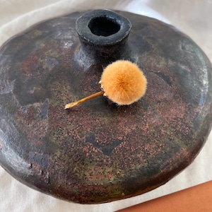 Könnte beinhalten: Eine handgefertigte, dunkelbraune Keramikvase mit strukturierter Oberfläche und einer kleinen, zylindrischen Öffnung. Ein kleiner, orangefarbener Pompon liegt auf der Vase. Die Vase hat eine rustikale, erdige Ästhetik.