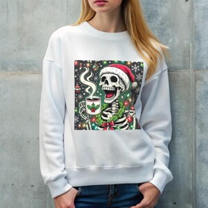 May include: White sweatshirt featuring a colorful graphic of a skeleton wearing a Santa hat, holding a mug with holly, and surrounded by Christmas lights and ornaments. The skeleton has a green wreath around its neck.