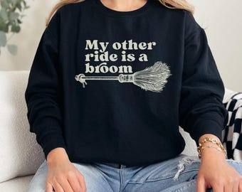 Może przedstawiać: Czarna bluza z okrągłym dekoltem z napisem "My other ride is a broom" i białą grafiką miotły. Osoba ma na sobie niebieskie dżinsy.