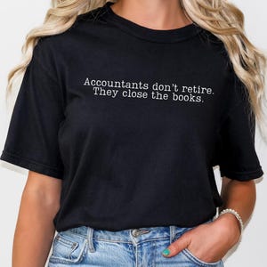a woman wearing a black comfort colors unisex tshirt. The minimalist graphic on the front reads in a white typewriter font "accountants don't retire. they close the books"