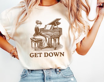 Puede incluir: Una camiseta de color crema con una ilustración vintage de una mujer tocando un piano de cola. El texto "GET DOWN" está impreso debajo de la ilustración.