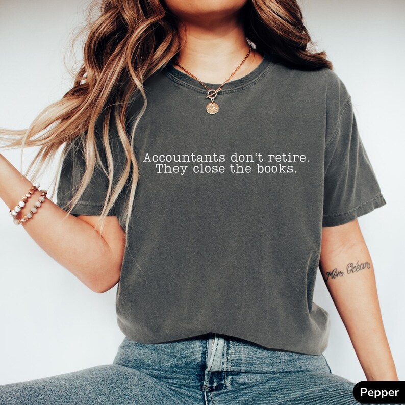 a woman wearing a pepper grey vintage wash comfort colors unisex t-shirt. The minimalist graphic on the front reads in a white typewriter font "accountants don't retire. they close the books"