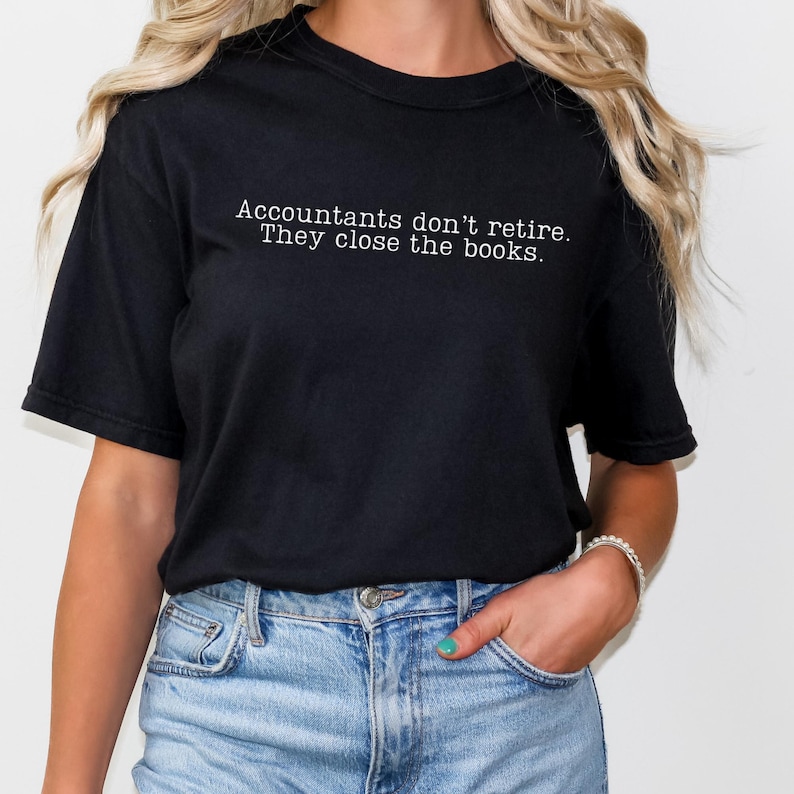 a woman wearing a black comfort colors unisex tshirt. The minimalist graphic on the front reads in a white typewriter font "accountants don't retire. they close the books"