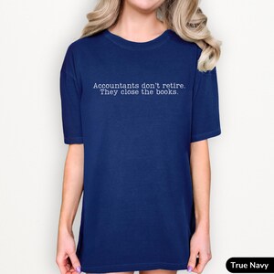 a young lady wearing a bright dark navy true blue comfort colors unisex t-shirt. The minimalist graphic on the front reads in a white typewriter font "accountants don't retire. they close the books"