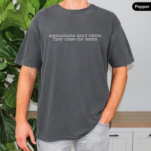 a man wearing a pepper dark gray vintage wash comfort colors unisex shirt. The minimalist graphic on the front reads in a white typewriter font "accountants don't retire. they close the books"