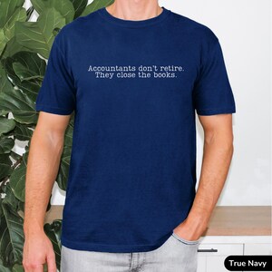 a young man wearing a bright dark navy true blue comfort colors unisex tee. The minimalist graphic on the front reads in a white typewriter font "accountants don't retire. they close the books"