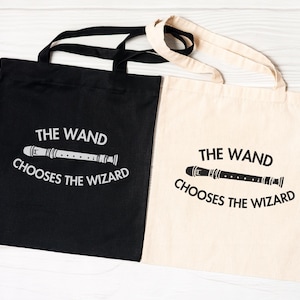 May include: Two tote bags, one black and one off-white, each featuring a graphic of a recorder instrument centred between the text "THE WAND" above and "CHOOSES THE WIZARD" below. The black bag has white text and graphics, while the off-white bag has black text and graphics.