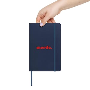 Könnte beinhalten: Ein marineblaues Notizbuch mit dem roten Wort "merde." auf der Vorderseite. Es hat einen blauen Gummibandverschluss und ein blaues Leseband. Das Notizbuch wird von einer Hand gehalten.