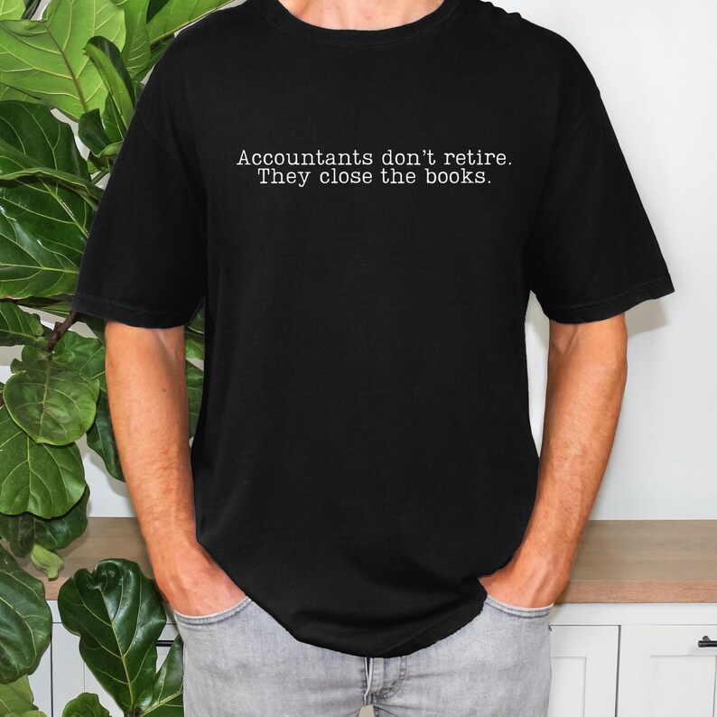 a guy wearing a black comfort colors unisex shirt. The minimalist graphic on the front reads in a white typewriter font "accountants don't retire. they close the books"