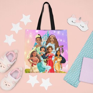 Pu&ograve; includere: Una borsa tote rosa e viola con un manico nero. La borsa presenta un'illustrazione colorata di sei principesse Disney, tra cui Tiana, Pocahontas, Mulan e Moana. Le principesse sono in piedi davanti a un castello rosa e viola con stelle sullo sfondo.