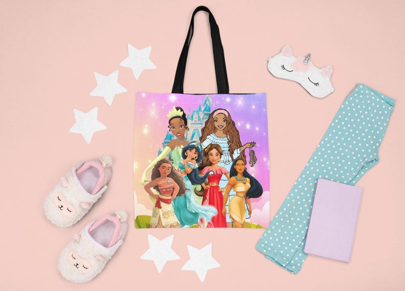 Pu&ograve; includere: Una borsa tote rosa e viola con un manico nero. La borsa presenta un'illustrazione colorata di sei principesse Disney, tra cui Tiana, Pocahontas, Mulan e Moana. Le principesse sono in piedi davanti a un castello rosa e viola con stelle sullo sfondo.