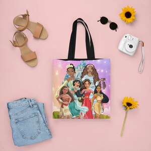 Pu&ograve; includere: Una borsa tote nera con una stampa colorata di principesse Disney. Le principesse sono in piedi davanti a uno sfondo rosa e viola con stelle e un castello. La borsa ha manici neri.