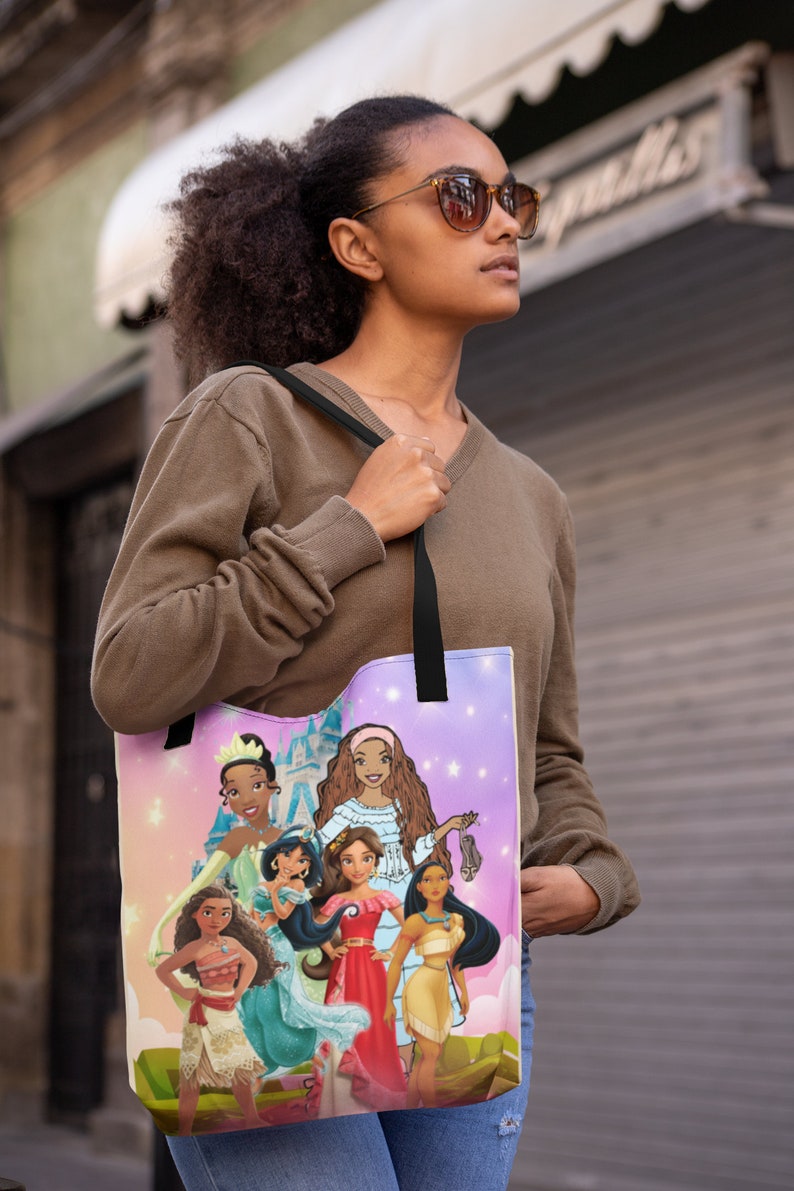 Pu&ograve; includere: Una borsa tote con sfondo rosa e viola che presenta un gruppo di principesse Disney. Le principesse indossano tutte abiti diversi e hanno acconciature diverse. La borsa ha spalline nere.