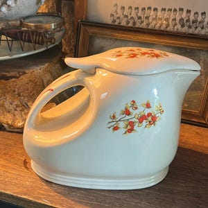 Könnte beinhalten: Cremefarbener Keramikkrug mit Deckel, mit einem großen Griff und einem Blumendesign mit roten und gelben Beeren. Der Krug wird auf einem Holzregal ausgestellt und zeigt eine Vintage-Ästhetik.