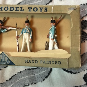 May include: Vintage model toy soldiers in a cardboard box. The hand-painted figures wear blue and white uniforms. The box reads "MODEL TOYS" and "HAND PAINTED".