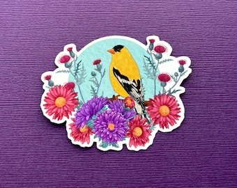Puede incluir: Un pájaro amarillo y negro con la cabeza blanca se posa en una rama rodeado de flores rosas, moradas y azules. El pájaro está posado en una rama con un fondo azul.
