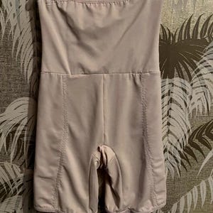 May include: A light beige, high-waisted shapewear garment with a strapless design. The garment features a smooth, seamless fabric with vertical seams and short legs. It is designed to provide body shaping and smoothing.