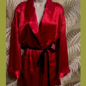 May include: A bright red satin robe featuring a shawl collar, long sleeves with folded cuffs, and a black velvet tie belt at the waist. The garment has a smooth, reflective finish and is displayed on a mannequin form, showcasing its structure.