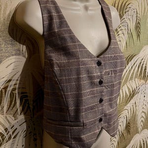 May include: A brown plaid vest with a V-neck and four black buttons. The vest has a tailored fit and a classic design. The fabric has a subtle pattern of brown, beige, and white lines. The vest is displayed on a mannequin.
