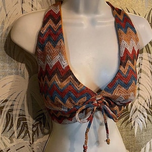 May include: A halter top with a chevron pattern in shades of red, orange, blue, and white. The top is displayed on a mannequin and features a tie closure at the front. The fabric appears to be knitted or crocheted.