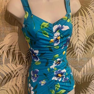 May include: A turquoise swimsuit with a floral pattern of white, blue, and yellow flowers and green leaves. The swimsuit has a ruched front and wide shoulder straps. The swimsuit is displayed on a mannequin.