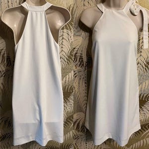 May include: Two white dresses on mannequins. The dresses are sleeveless with a halter neckline. One dress has a bow detail on the shoulder. The dresses are displayed against a patterned background.