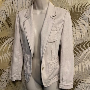 May include: A light gray blazer with a herringbone pattern. The blazer has a notched lapel, two front pockets, and a single-button closure. Dark stitching outlines the seams and pockets. The jacket is displayed on a mannequin.