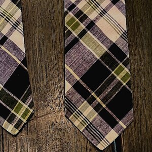 May include: A necktie featuring a plaid pattern with intersecting lines in shades of black, olive green, light purple, and cream. The fabric displays a woven texture with a pointed end, showcasing a classic grid design across the entire surface.