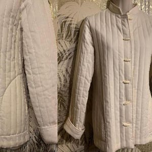 May include: Cream-colored quilted jacket with a mandarin collar and long sleeves. The jacket features vertical stitching and is fastened with four small, round buttons. The cuffs are turned up, revealing a lighter shade of the same color.