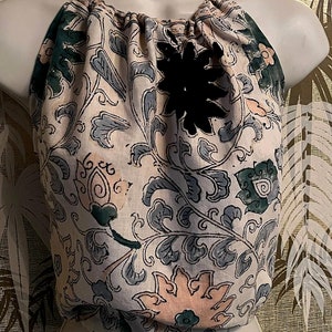 May include: A cream-coloured halter top with a floral design in green and blue. It has a black flower-shaped detail and a drawstring neckline. The fabric appears soft and textured, possibly linen or cotton.