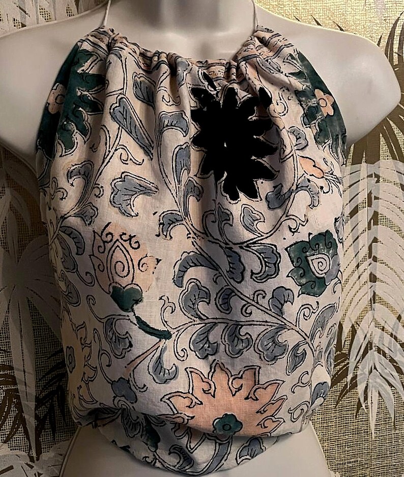 May include: A cream-coloured halter top with a floral design in green and blue. It has a black flower-shaped detail and a drawstring neckline. The fabric appears soft and textured, possibly linen or cotton.