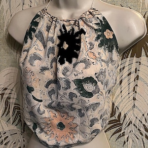 May include: A halter top with a floral design in white, grey, green, and peach. It has a black flower appliqu&eacute; and a halter neck. The fabric has a soft, slightly wrinkled texture. The top is displayed on a mannequin.