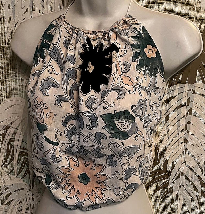 May include: A halter top with a floral design in white, grey, green, and peach. It has a black flower appliqu&eacute; and a halter neck. The fabric has a soft, slightly wrinkled texture. The top is displayed on a mannequin.