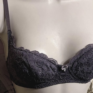 May include: A dark gray bra with intricate lace detailing. The bra features adjustable straps and a small bow accent between the cups. The lace pattern is floral, and the bra appears to be unworn.