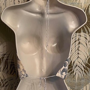 May include: A halter top with a blue and white floral pattern, shown on a white mannequin. The top is tied at the neck and back with white string. The background features a wallpaper with a leaf design.