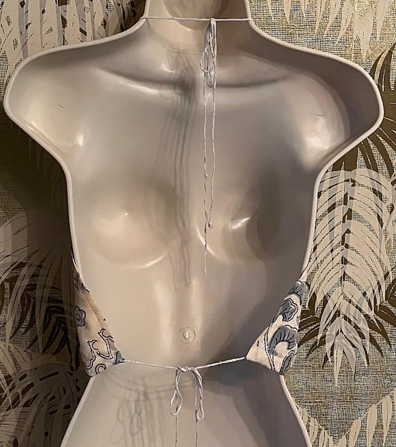 May include: A halter top with a blue and white floral pattern, shown on a white mannequin. The top is tied at the neck and back with white string. The background features a wallpaper with a leaf design.