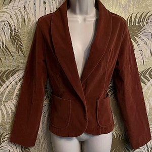 May include: A rust-colored blazer with a shawl collar and two front pockets. The blazer is displayed on a mannequin. The fabric appears to be a soft, velvety material. The blazer has a single-button closure.